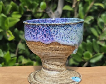 May include: A handmade ceramic goblet with a pedestal base. The rim is a blend of blue and purple hues, transitioning to a tan midsection and a textured, brown base. The goblet is displayed on a wooden surface.