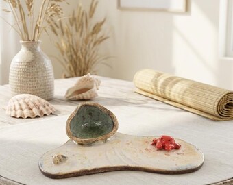 May include: A ceramic serving tray with an irregular, organic shape featuring a sandy-colored textured surface. The tray includes a small, attached green bowl, a tiny sculpted red crab, and a small seashell accent on the flat base.