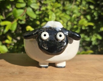 May include: A ceramic sheep figurine with a white body, black face, and small white eyes. The sheep has a glossy finish and is standing on four small legs. The background is blurred green foliage.