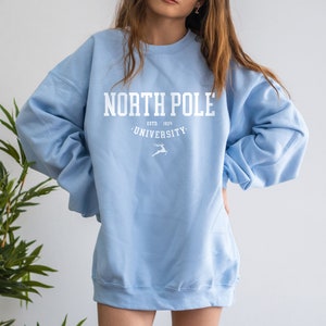May include: A light blue sweatshirt with the text "North Pole University" in white. The text is surrounded by a white circle with the words "Estd. 1824" above the university name and a white reindeer silhouette below.
