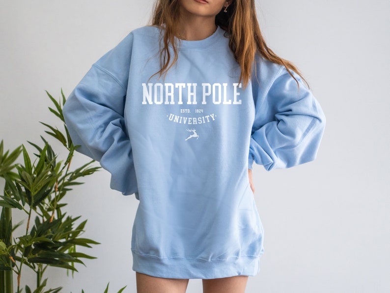 May include: A light blue sweatshirt with the text "North Pole University" in white. The text is surrounded by a white circle with the words "Estd. 1824" above the university name and a white reindeer silhouette below.