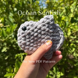 May include: A crocheted gray sunfish with a black eye is held in a hand. The text "Ocean Sunfish" and "No-Sew PDF Pattern" are visible. The sunfish is made of a soft, textured yarn.