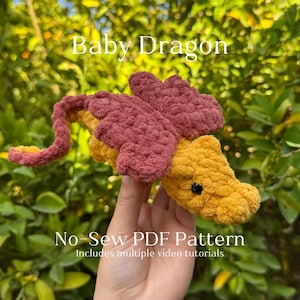 May include: A crocheted baby dragon plush toy in pink and yellow. The dragon is being held in a hand and the text "Baby Dragon No-Sew PDF Pattern Includes multiple video tutorials" is visible below the dragon.