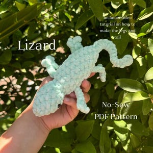 Op de afbeelding: Een lichtgroen gehaakt speelgoed-hagedis. Het speelgoed wordt in een hand gehouden. De afbeelding bevat de tekst "Lizard", "Includes video tutorial on how to make the legs and tail", "No-Sew" en "PDF Pattern".