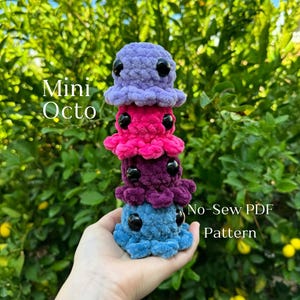 Könnte beinhalten: Ein Stapel von vier kleinen gehäkelten Oktopus-Plüschtieren in Lila, Rosa, Dunkelviolett und Blau. Die Plüschtiere sind übereinander gestapelt und werden in einer Hand gehalten. Der Text "Mini Octo No-Sew PDF Pattern" ist im Bild sichtbar.