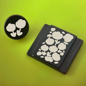 Könnte beinhalten: Ein schwarzer runder Knopf und ein schwarzer quadratischer Magnet mit weißem Blumendesign auf einem limettengrünen Hintergrund. Der Knopf zeigt zwei Blumen, der Magnet mehrere.