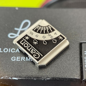 May include: A square silver-toned metal pin featuring a black enamel inlay with the Canon logo. The design includes a graphic representation of a light meter with aperture numbers 4, 5.6, 8, 11, and 16, alongside weather icons for sun and clouds.