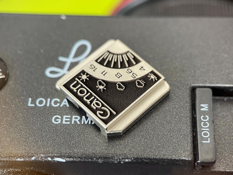 May include: A square silver-toned metal pin featuring a black enamel inlay with the Canon logo. The design includes a graphic representation of a light meter with aperture numbers 4, 5.6, 8, 11, and 16, alongside weather icons for sun and clouds.