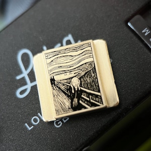 Op de afbeelding: Een rechthoekige, goudkleurige pin met een zwart-witte afbeelding van Edvard Munchs kunstwerk "De Schreeuw". De pin ligt op een donkergrijze ondergrond, met een gedeeltelijk zicht op een camerabody en het Leica-logo.