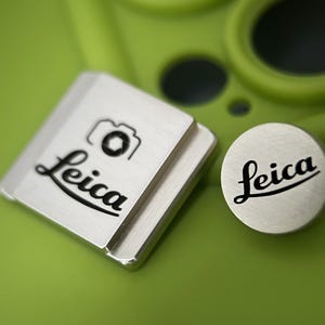 Könnte beinhalten: Silbernes Leica Kamera Zubehör auf grünem Untergrund. Das quadratische Teil hat ein Kamera-Symbol und das Leica-Logo. Ein rundes Teil mit dem Leica-Logo ist ebenfalls sichtbar. Der Hintergrund ist verschwommen grün.