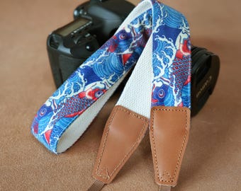 May include: A camera strap with a blue and white wave and fish pattern. The strap has a white canvas center and brown leather ends. A black camera is in the background.