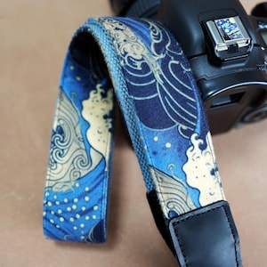 Peut inclure: Un sangle de caméra bleu et blanc avec un motif de vagues. La sangle est en tissu et possède une sangle de réglage en cuir noir.