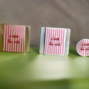K&ouml;nnte beinhalten: Drei Metallobjekte mit dem Schriftzug "c'est la vie" in roter Schrift. Die Objekte haben ein rosa-wei&szlig; gestreiftes Design. Die Objekte sind quadratisch, rechteckig und kreisf&ouml;rmig.