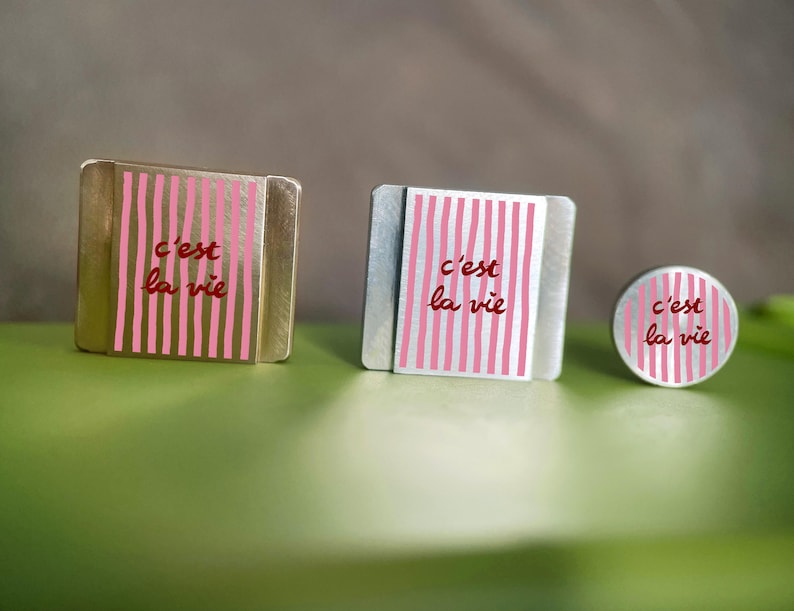 K&ouml;nnte beinhalten: Drei Metallobjekte mit dem Schriftzug "c'est la vie" in roter Schrift. Die Objekte haben ein rosa-wei&szlig; gestreiftes Design. Die Objekte sind quadratisch, rechteckig und kreisf&ouml;rmig.