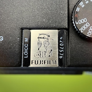 May include: A black camera body featuring a silver hot shoe cover engraved with a cat holding a sword in its mouth and the text FUJIFILM. The camera includes a shutter speed dial with white numbers and the text LEICA CAMERA AG visible nearby.