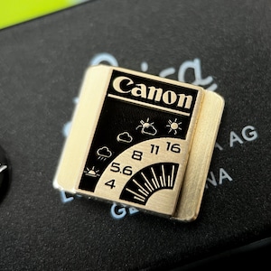 Könnte beinhalten: Ein goldfarbenes Canon-Zubehör mit schwarzer Oberfläche, das Wettersymbole, Zahlen und ein Sonnenstrahl-Design zeigt. Das Canon-Logo ist oben prominent platziert. Das Zubehör ist rechteckig mit abgerundeten Kanten.