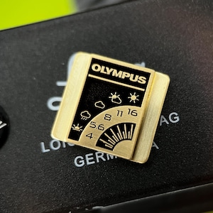 May include: A gold-coloured Olympus camera accessory with black text and graphics. The accessory features the Olympus logo and weather symbols. Text includes numbers and the word "GERMANY".