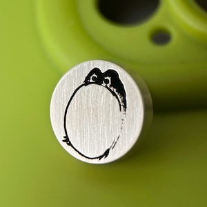 Op de afbeelding: Een ronde, zilveren stempel met een zwart pinguïnontwerp. De pinguïn is afgebeeld in een eenvoudige, minimalistische stijl. De stempel ligt op een heldergroen oppervlak, met een wazig groen object op de achtergrond.