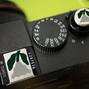 May include: A black camera body with a silver and green floral graphic. The camera has a dial with white numbers and a black control knob. The graphic is a stylized flower. The text "LOICC M" and "4207576" are visible.