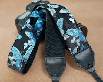 May include: A black camera strap with a repeating pattern of blue and white butterflies. The strap has a black canvas center and black leather ends. The camera strap is designed to be attached to a camera.