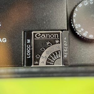 May include: A black metal camera accessory featuring a silver rectangular plate with the Canon logo and a weather-based exposure guide. The guide displays icons for various lighting conditions alongside aperture settings 4, 5.6, 8, 11, and 16.