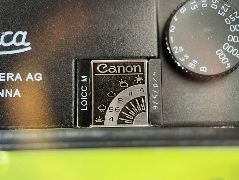 May include: A black metal camera accessory featuring a silver rectangular plate with the Canon logo and a weather-based exposure guide. The guide displays icons for various lighting conditions alongside aperture settings 4, 5.6, 8, 11, and 16.
