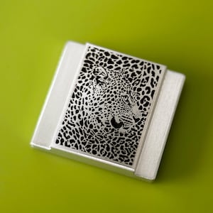 May include: A silver-toned rectangular brooch with a detailed black and white leopard design. The leopard's face is centered on the brooch, set against a bright green background. The brooch has a brushed metal finish.