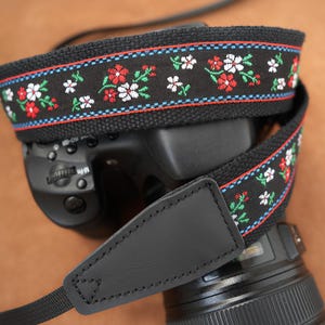 Könnte beinhalten: Schwarzer Kameragurt mit floralem Design. Der Gurt hat rote, weiße und grüne Blumen mit blau-roter Verzierung. Der Gurt ist an einem schwarzen Kameragehäuse mit einem schwarzen Lederriemen befestigt.