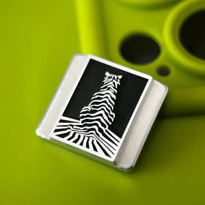 Könnte beinhalten: Eine quadratische Anstecknadel mit silbernem Rand und schwarzem Zentrum. Die Anstecknadel zeigt ein weißes Zebra-Design mit einem ausgeprägten wellenförmigen Muster. Die Anstecknadel befindet sich auf einem hellgrünen Hintergrund.