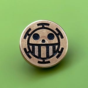 May include: A gold-colored metal pin with a black and white skull and crossbones design. The skull has a single tooth and two eyes. The design is surrounded by a circle of black lines.