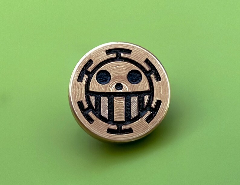 May include: A gold-colored metal pin with a black and white skull and crossbones design. The skull has a single tooth and two eyes. The design is surrounded by a circle of black lines.