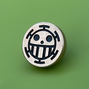 May include: A silver-colored metal pin with a black and white skull and crossbones design. The skull has two black eyes and a white mouth with teeth.