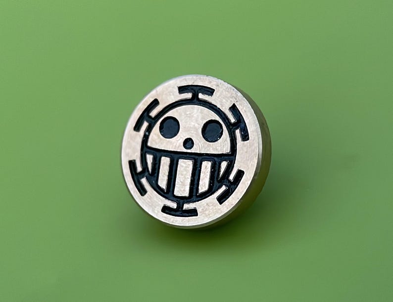 May include: A silver-colored metal pin with a black and white skull and crossbones design. The skull has two black eyes and a white mouth with teeth.
