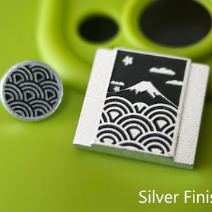 May include: Two silver-toned metal pins. One is round with a black wave pattern. The other is rectangular with a black and white mountain and wave design. The text "Silver Finish" is at the bottom of the image.