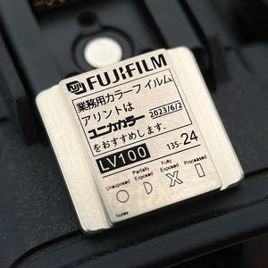 May include: A silver metal film canister with the text "FUJIFILM 業務用カラーフイルム アリントは ユニカカラー 2023/6/2 をおすすめします。 LV100 135-24" printed on it. The canister has a label with the text "Partially Unexposed Fully Exposed Processed ODXI Noles".