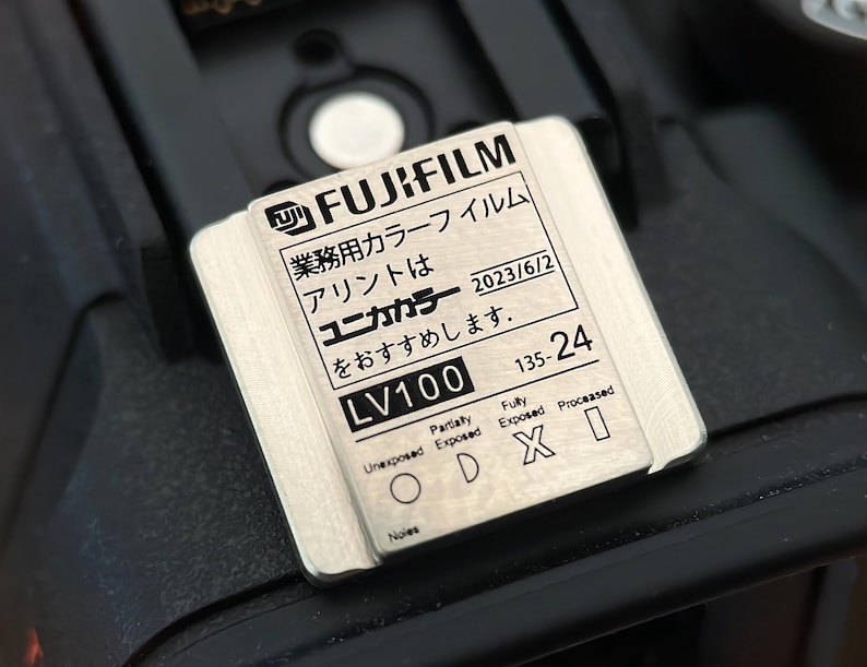 May include: A silver metal film canister with the text "FUJIFILM 業務用カラーフイルム アリントは ユニカカラー 2023/6/2 をおすすめします。 LV100 135-24" printed on it. The canister has a label with the text "Partially Unexposed Fully Exposed Processed ODXI Noles".