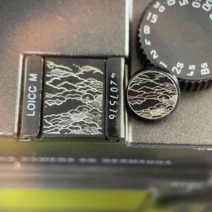 Puede incluir: Un conjunto de accesorios de cámara en color negro que incluye una placa cuadrada y un botón circular, ambos grabados con un patrón blanco de nubes y olas. La placa muestra el texto LOICC M y 4207576 junto a un dial de la cámara.