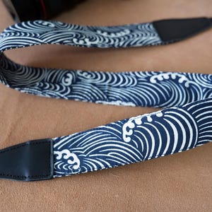 May include: A navy blue and white patterned camera strap. The strap features a repeating wave design. The ends of the strap are black leather. The strap is designed to be worn around the neck.