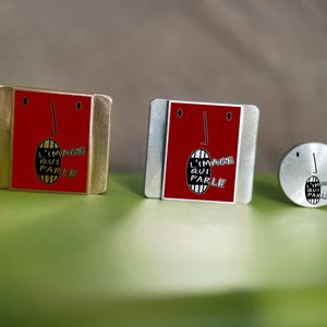 May include: Two rectangular metal pins and one circular metal pin, each featuring a red face graphic with a black line nose, two eyes, and an open mouth containing the text L'IMAGE QUI PARLE in black and white lettering on a striped background.