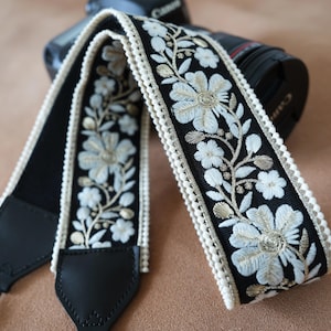 May include: Black camera strap with embroidered floral design in white and gold. The strap has a cream-colored trim and black leather ends. The strap is designed to attach to a camera.