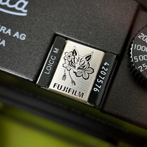 Może przedstawiać: Zbliżenie czarnego aparatu z srebrną płytką z wystylizowaną twarzą lisa z kwiatowymi akcentami. Płytka jest oznaczona napisem "FUJIFILM" i "4207576". Po prawej stronie widoczna jest tarcza z numerami.