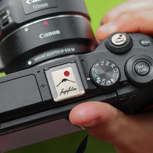 May include: A black Canon camera with a lens attached. The camera has a dial with the letters "M-Fn", "ON", and "OFF". The camera also has a sticker that says "fujifilm" with a mountain and a red sun.