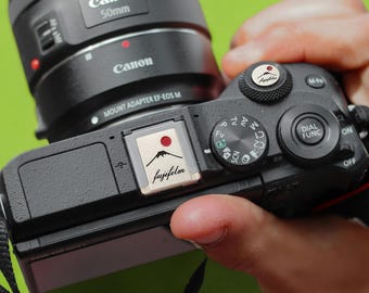 May include: A black Canon camera with a lens attached. The camera has a dial with the letters "M-Fn", "ON", and "OFF". The camera also has a sticker that says "fujifilm" with a mountain and a red sun.
