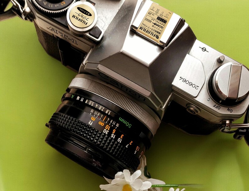May include: A silver Canon film camera with a 50mm lens. The camera has a black lens cap and a black strap. The camera is on a green surface.