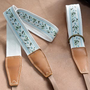 May include: A cream-colored camera strap with a floral embroidered design in shades of blue and green. The strap features brown leather ends with beige stitching and a metal ring for attachment. The strap is designed for a camera.