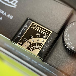 May include: A dark gray camera top plate featuring a metallic exposure guide plate with the Canon logo, weather icons, and aperture settings ranging from 4 to 16. A black shutter speed dial with white markings is positioned to the right of the guide.