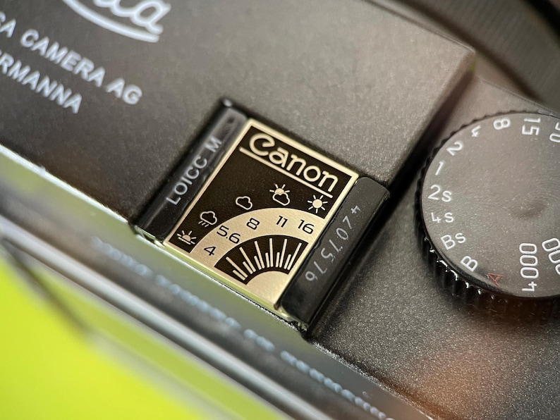 May include: A dark gray camera top plate featuring a metallic exposure guide plate with the Canon logo, weather icons, and aperture settings ranging from 4 to 16. A black shutter speed dial with white markings is positioned to the right of the guide.