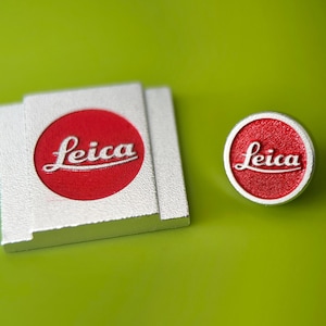 Op de afbeelding: Twee Leica logo items op een groene ondergrond. Eén is een rechthoekig zilverkleurig stuk met een rode cirkel en witte Leica tekst. De andere is een rond zilverkleurig stuk met dezelfde rode cirkel en witte Leica tekst.