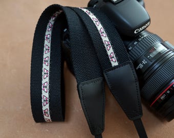 May include: Black camera strap with a white and pink butterfly pattern. The strap is made of durable fabric and features black leather ends. A Canon camera with a lens is visible in the background.
