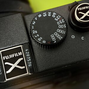 Könnte beinhalten: Nahaufnahme eines schwarzen Fujifilm Kameragehäuses. Die Oberseite der Kamera verfügt über ein Rad mit Zahlen und Einstellungen, ein silbernes und schwarzes Fujifilm-Logo und ein Modusrad. Die Kamera hat eine strukturierte Oberfläche und eine Seriennummer.